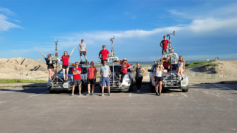 Students stand on and in front of three cars modified with special data collection equipment.